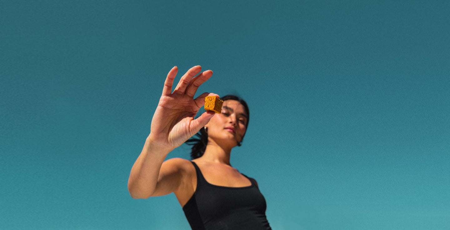 A woman in a black tank top holding up a small golden-brown GEM Bite against a vibrant teal sky.