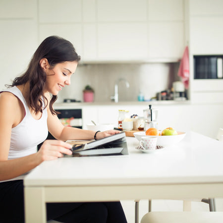 Young woman sitting at a white dining table while smiling, sipping herbal tea, and reading.
