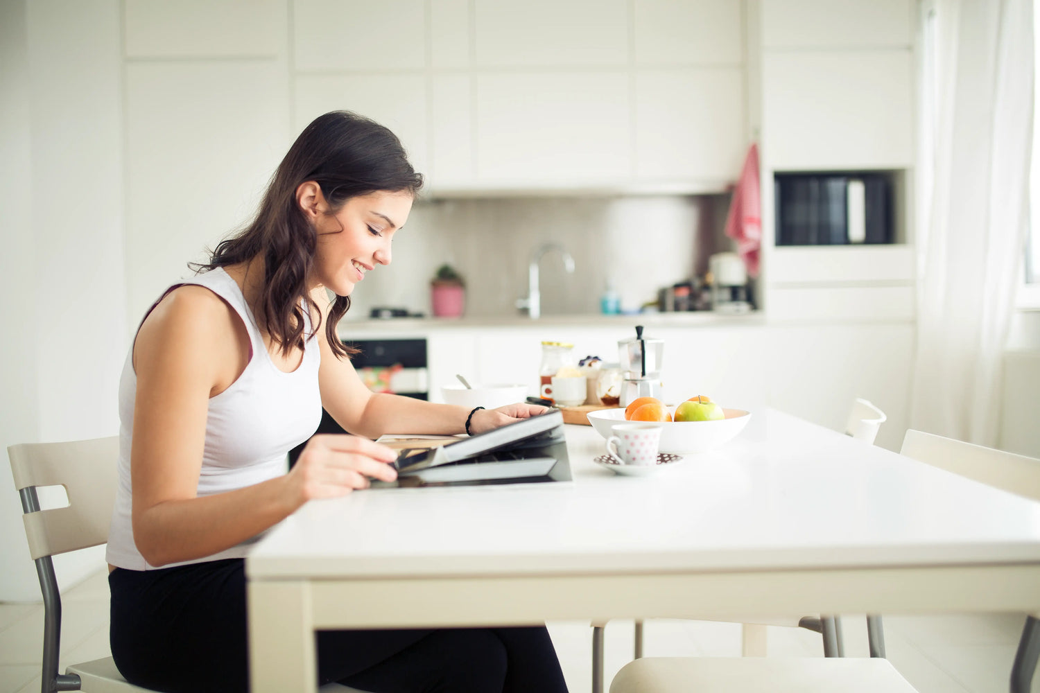 Young woman sitting at a white dining table while smiling, sipping herbal tea, and reading.